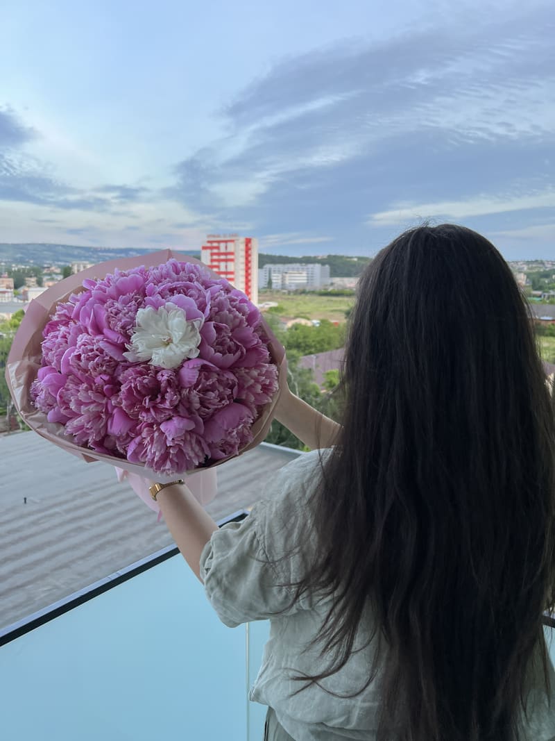 Femeie văzută din spate, ținând un buchet mare de bujori roz pe balcon